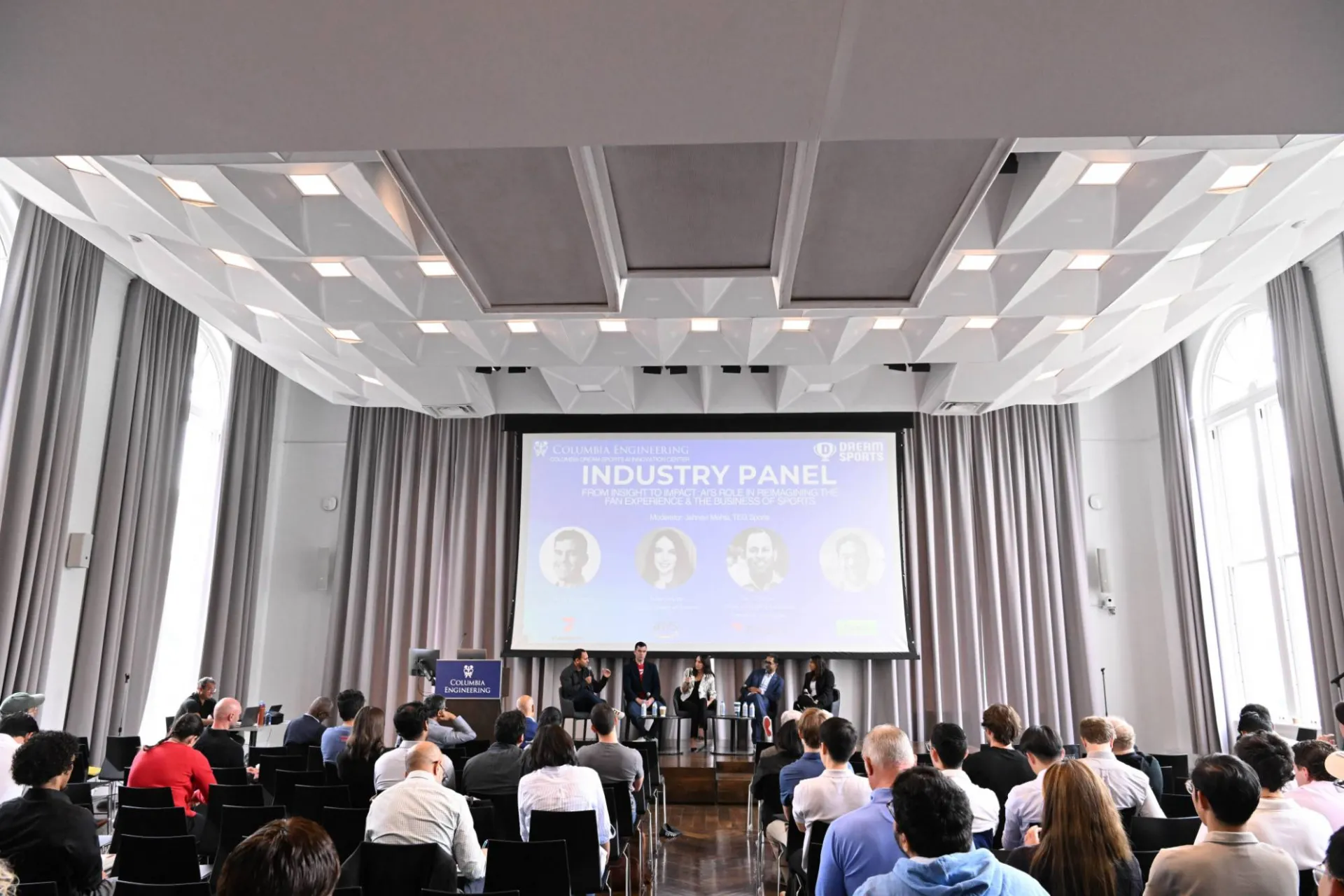 image of the back of the room, showing the audience and projection of "industry panel" graphic with 5 panelists sitting on the stage