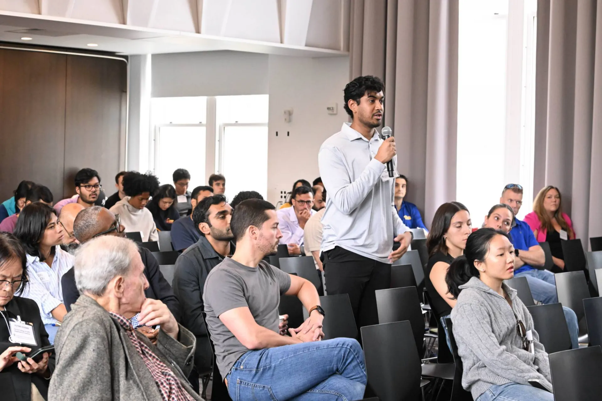image of audience member wearing a light grey blazer standing from the crowd, speaking into a handheld microphone
