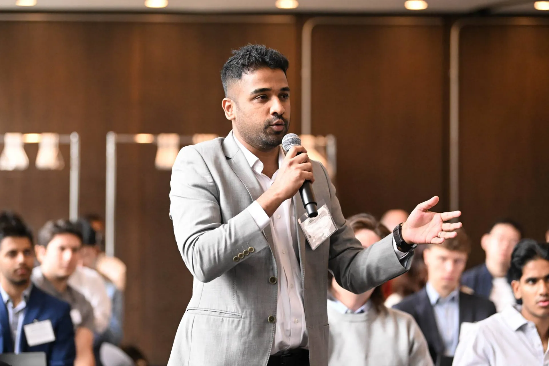 image of audience member wearing a light grey blazer standing from the crowd, speaking into a handheld microphone