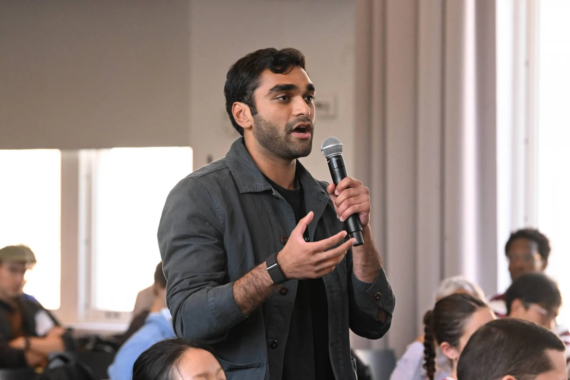 single audience member wearing a dark grey jacket standing from the crowd, speaking into a hand held microphone