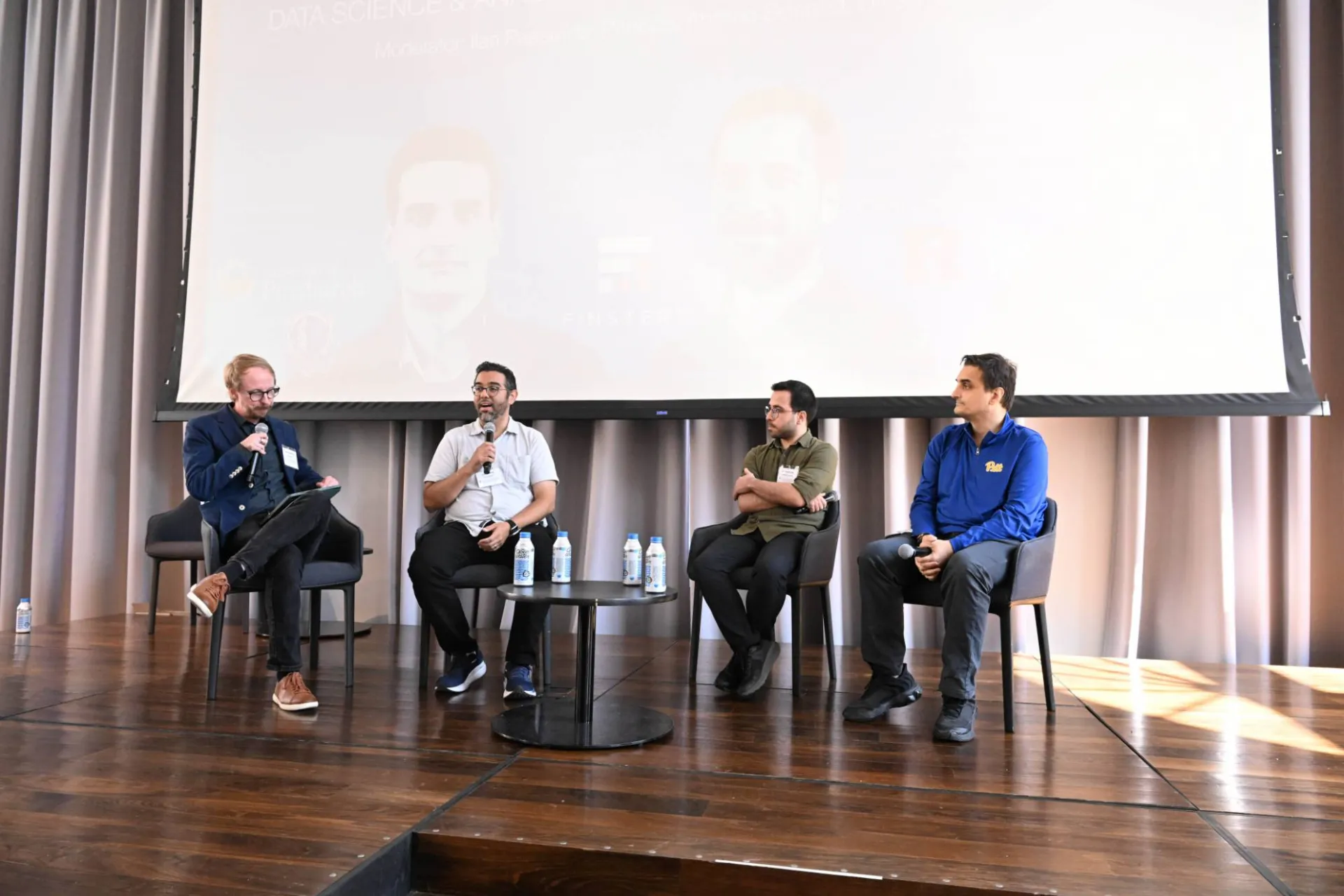 photo of 4 speakers on stage, the two on the left holding a microphone and addressing peers