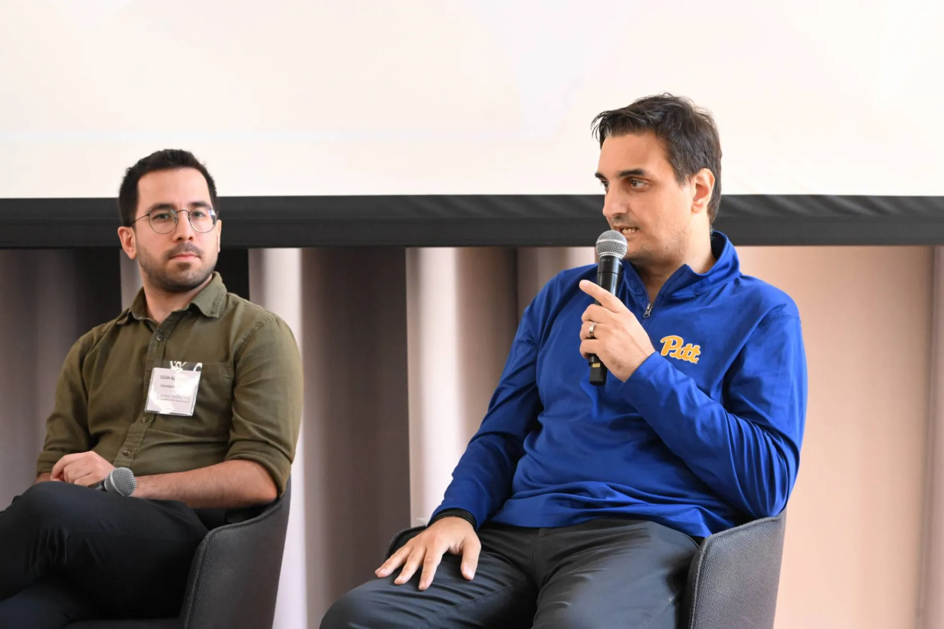close up of 2 speakers on stage, the one on the far right holding a microphone and addressing peer(s)