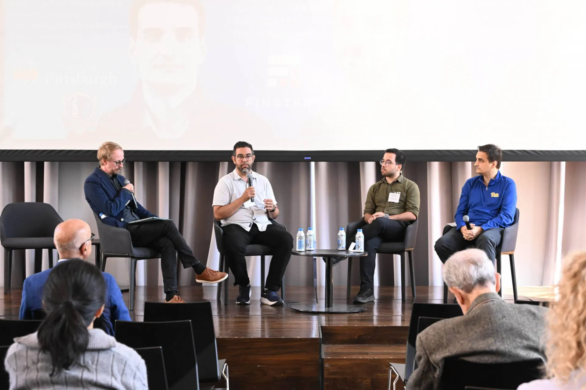 photo of 4 speakers sitting on stage, one addressing the audience with a microphone