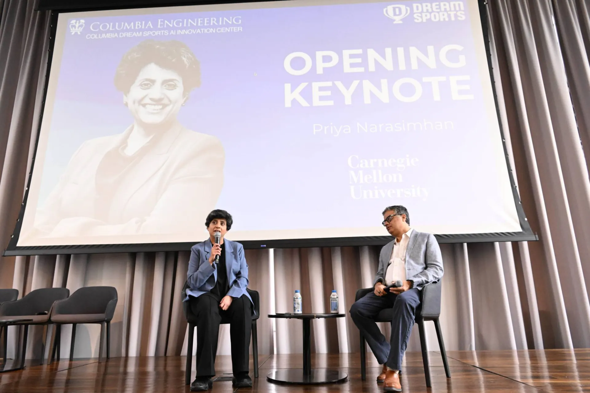 image of the opening keynote speaker, Priya Narasimhan onstage with facilitator, sitting in front of a projector displaying a graphic introducing the speaker