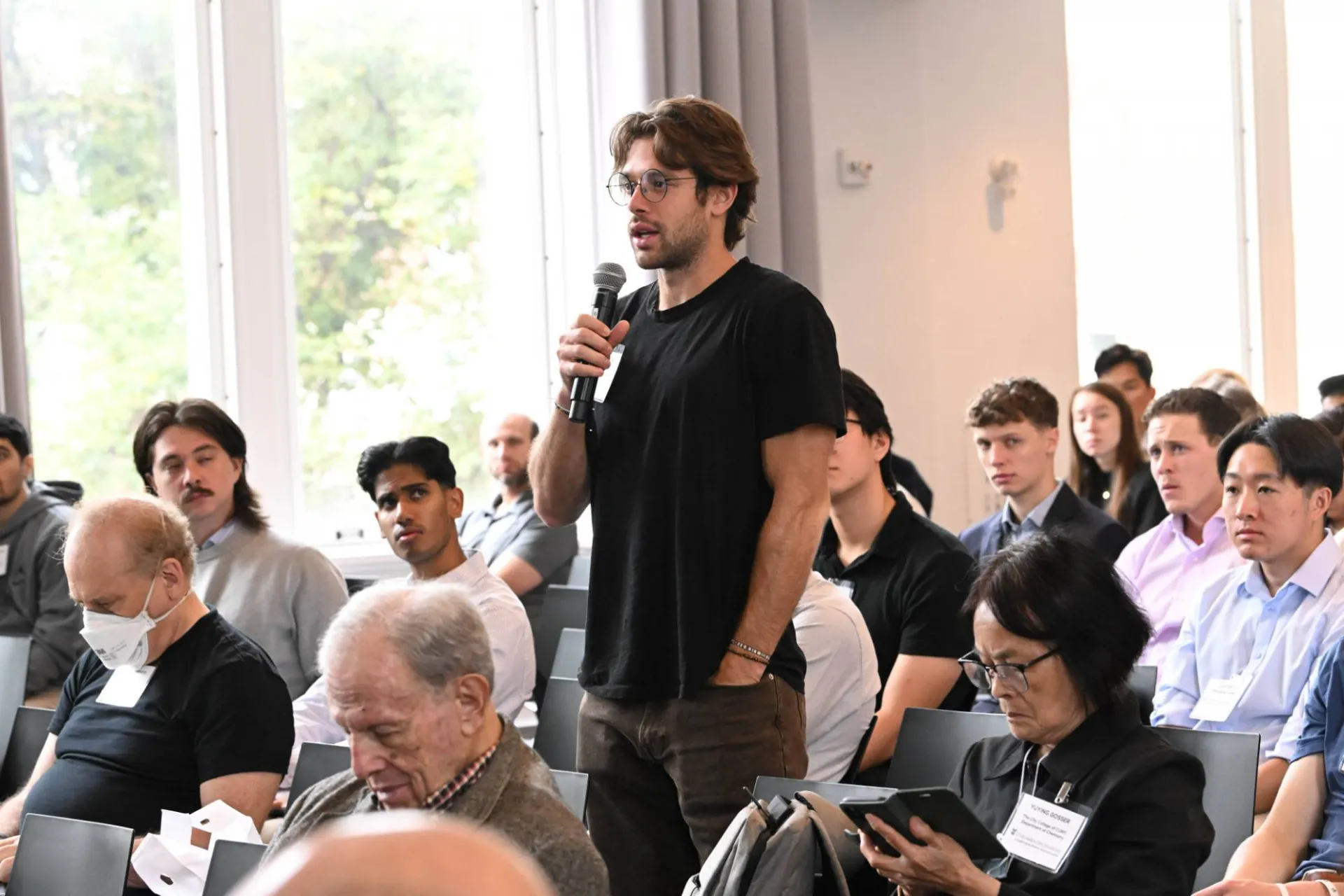 image of audience member wearing a black shirt standing up from the crowd, speaking into a microphone in hand