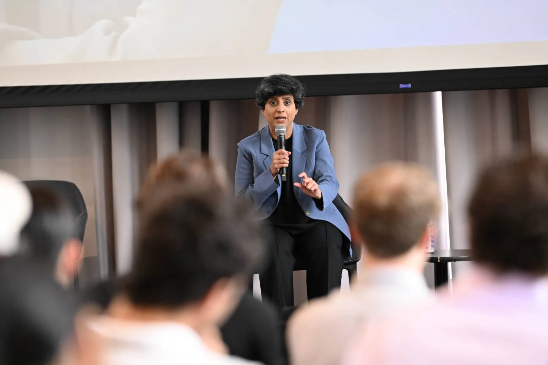 image of panelist wearing a blue blazer sitting on stage, addressing the audience while speaking into a microphone