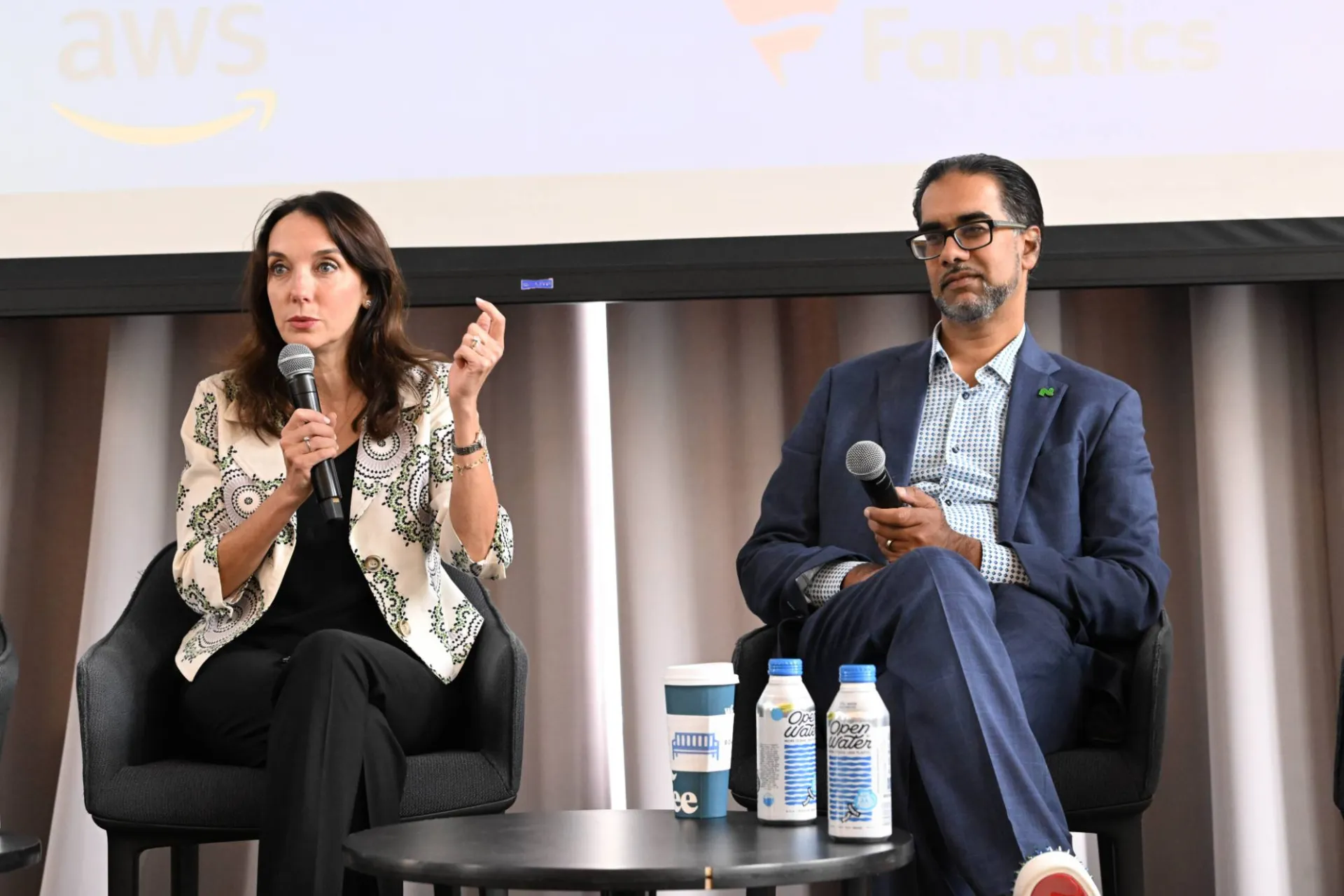 image of 2 panelists speaking on stage, the one on the left speaking into a microphone