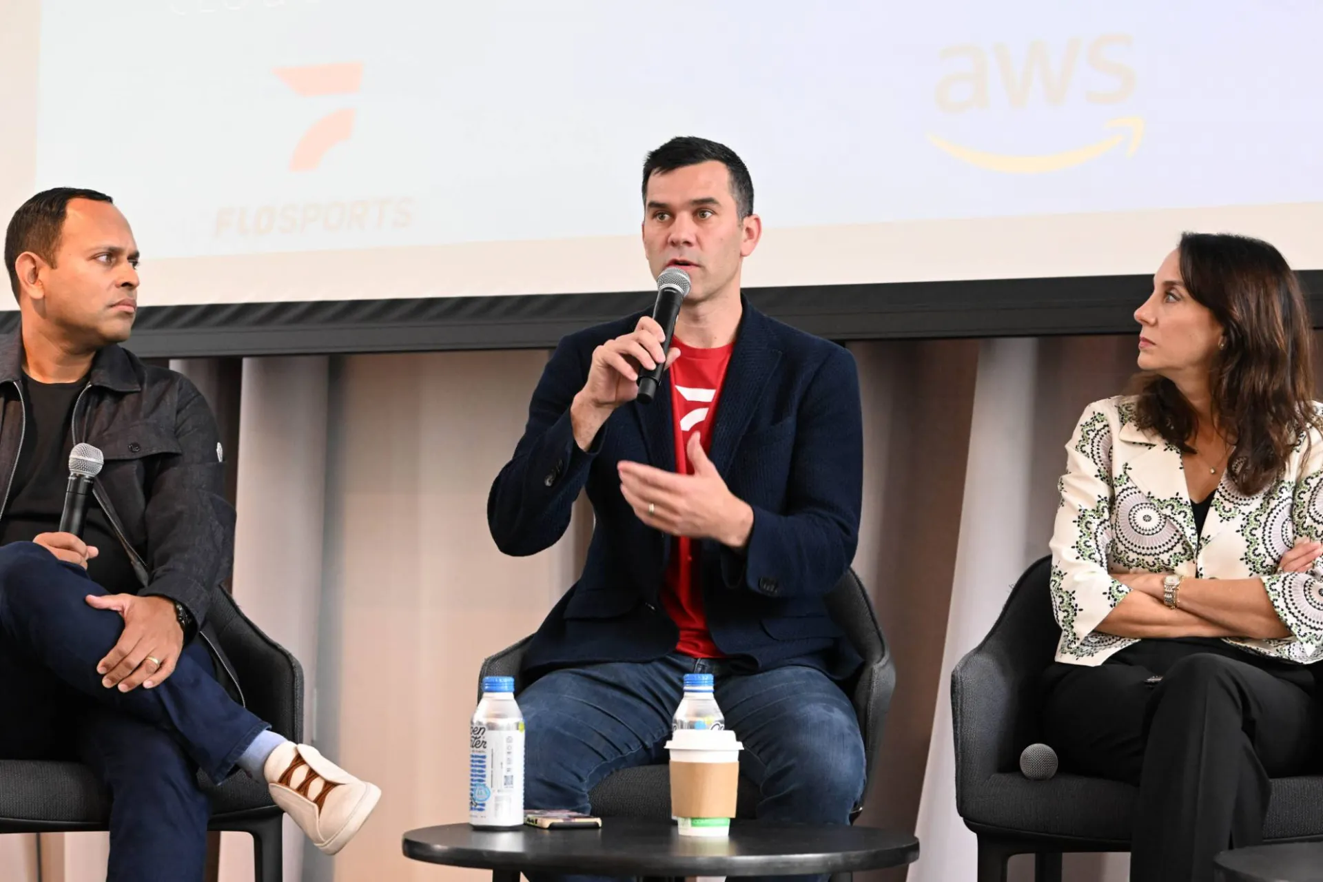 image of 3 panelists speaking on stage, the one in the middle speaking into a microphone while the other two look at them