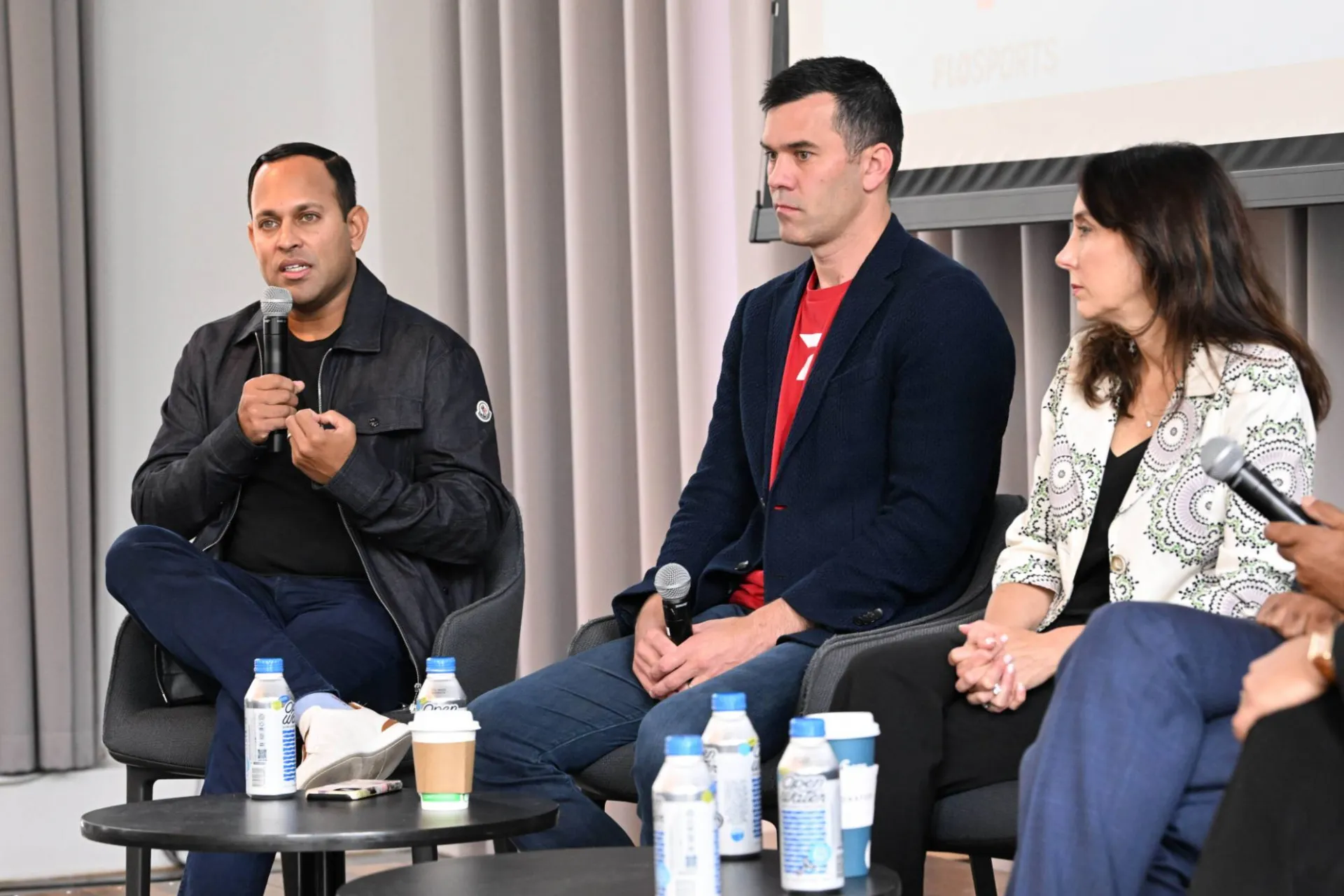 image of 3 panelists speaking on stage, one speaking into a microphone